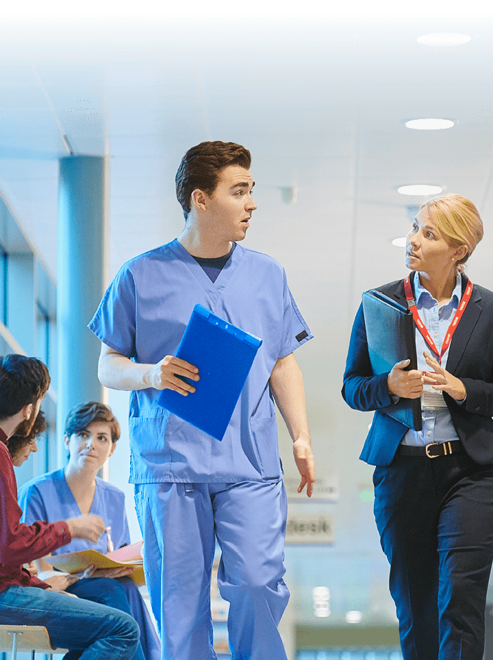 a mid adult male doctor speaks with a businesswoman , sales person on the hospital corridor as they head to a meeting ,with a young female nurse and patients in the busy corridor .