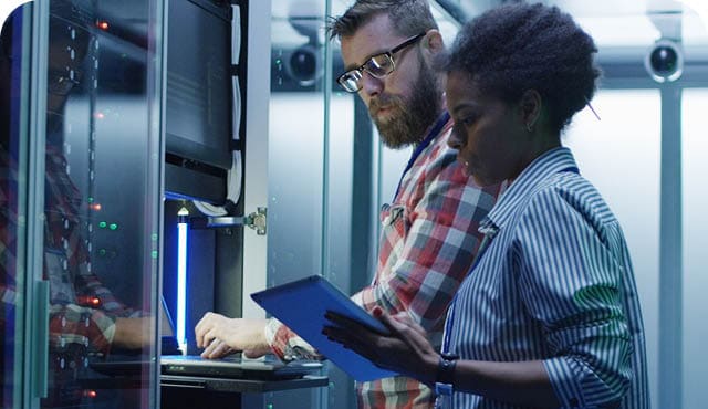 Modern multiethnic man and woman with tablet using laptop in server room while checking servers