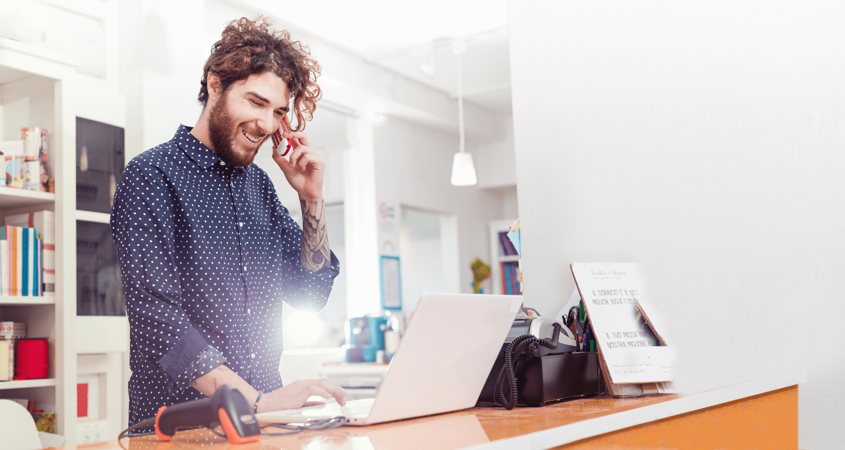 indoors, independent, shop, small business, working, standing, casual clothing, pois, shirt, beard, wireless technology, bookshop, books, curly hair, working, jeans, proud, smiling, toothy smile, laptop, computer barcode scanner, cash register, tattoos, holding, phone, mobile phone, typeing,