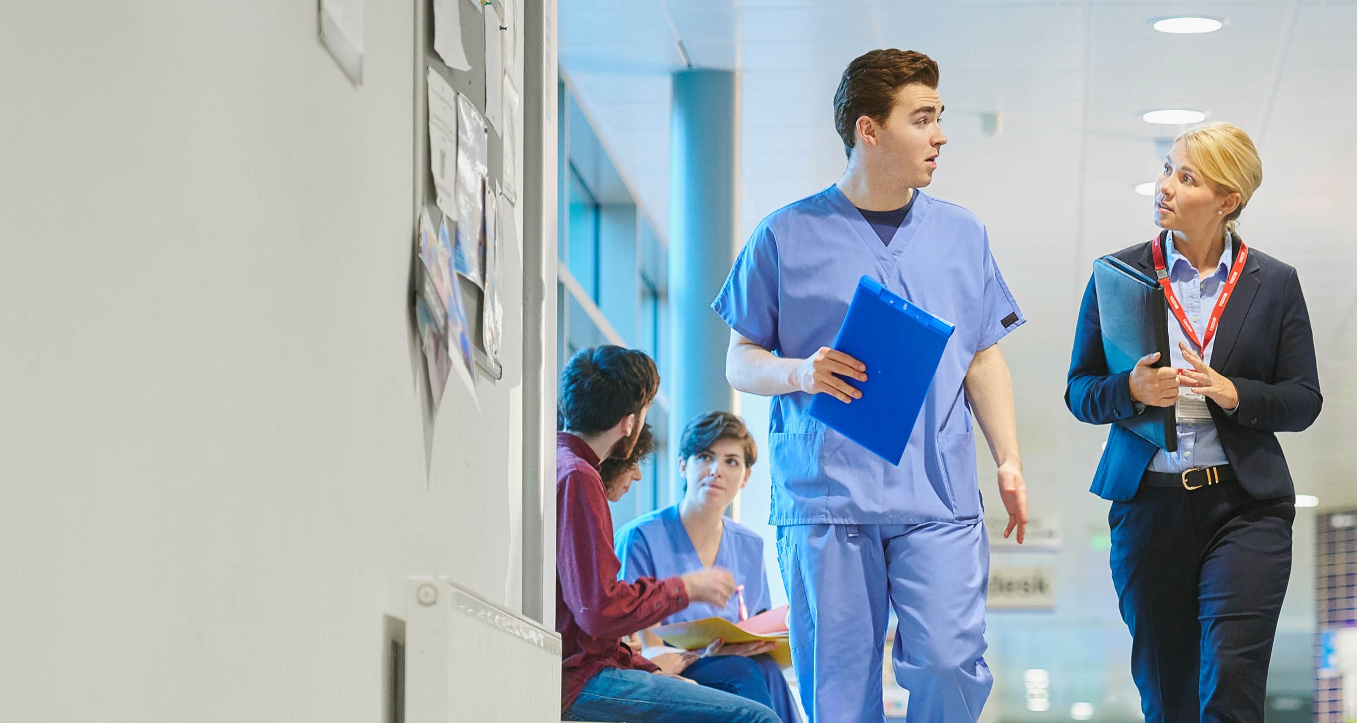 a mid adult male doctor speaks with a businesswoman , sales person on the hospital corridor as they head to a meeting ,with a young female nurse and patients in the busy corridor .